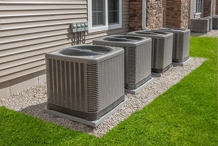 Four outdoor air conditioning units sit on gravel next to a building with siding and bricks, surrounded by lush green grass. Homeowners often wonder what size heat pump would best suit their space for optimal efficiency and comfort.